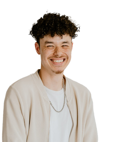 A young man with curly dark hair and a light goatee smiles while wearing a cream jacket, white shirt, and chain necklace against a plain white background.