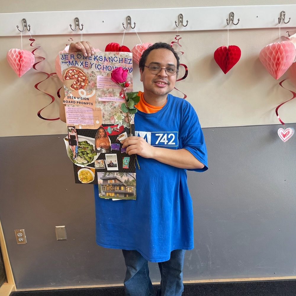 A man in a blue shirt holds a vision board with various images and text, standing in front of Valentine’s Day decorations on a gray and white wall.