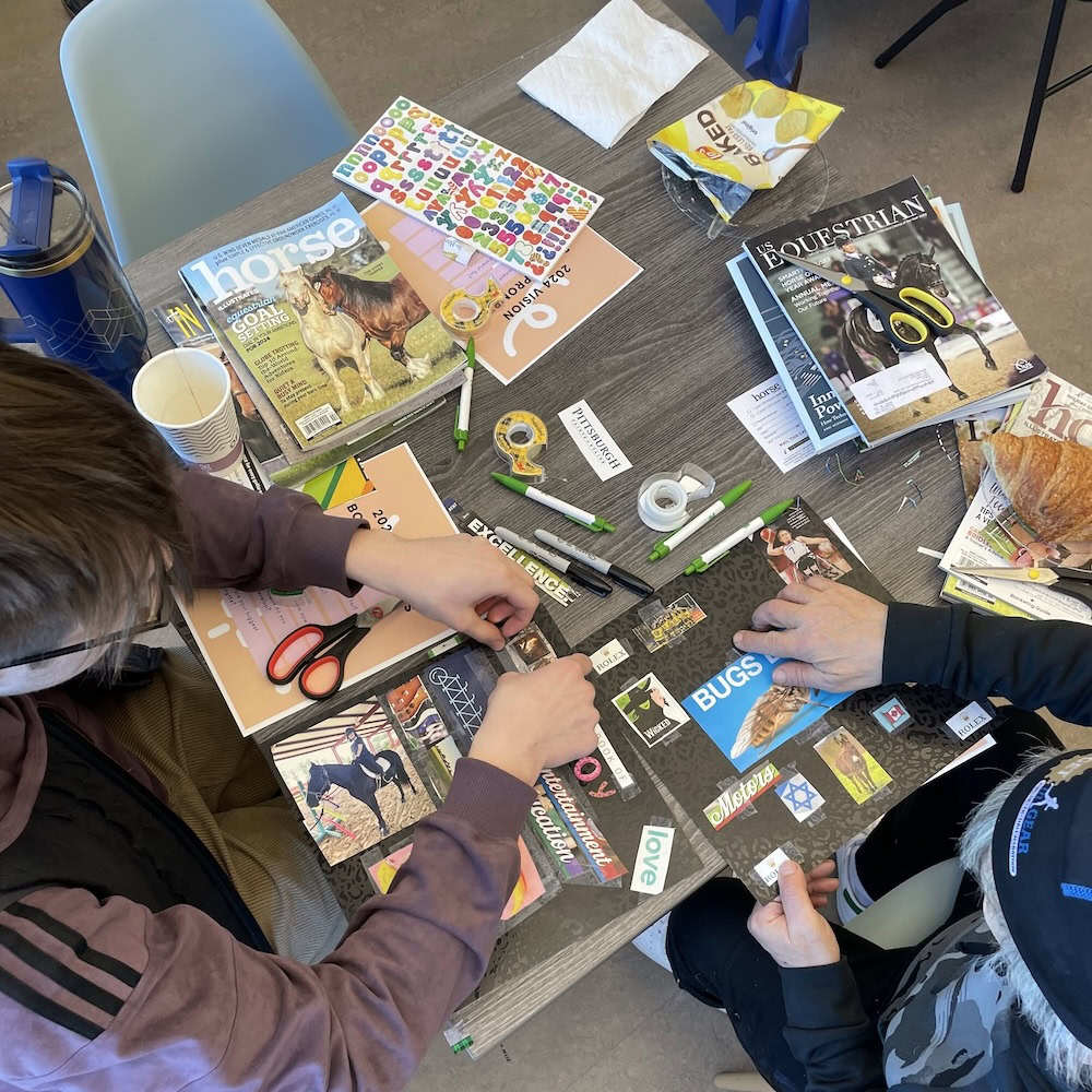 Two people work on a collage at a table, surrounded by magazines, scissors, tape, markers, stickers, and cups. The collage features cut-out words and images.