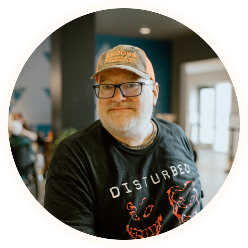 A man wearing glasses, a trucker hat, and a black "Disturbed" band t-shirt sits indoors, facing the camera with a slight smile—capturing a relaxed moment supported by Individualized Home Supports.