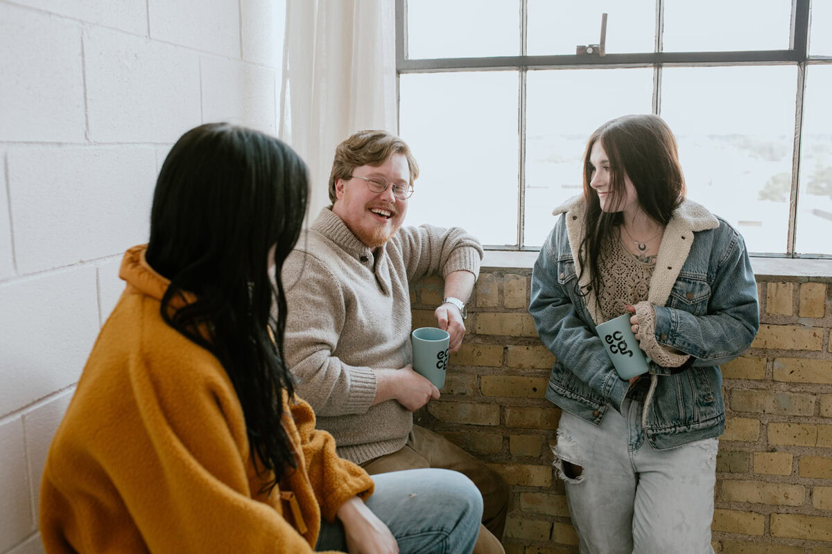 Three people sit and stand by a window, holding mugs and talking. Two women face a man who is smiling and seated on the windowsill.
