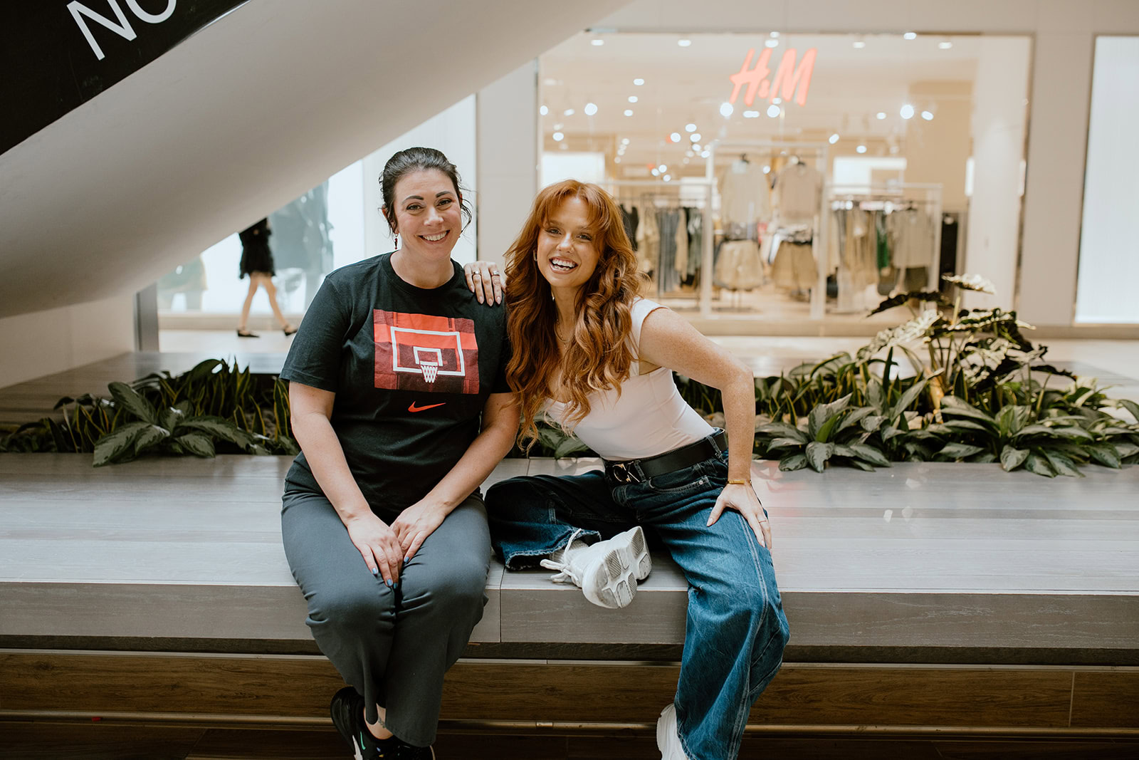 Two women sit side by side on a bench in a shopping mall, smiling at the camera. ECCG banners and clothing store displays are visible in the background.