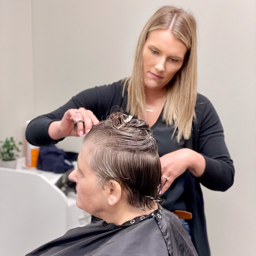 A hairdresser with straight blonde hair is cutting the short, wet hair of a seated client in a salon.