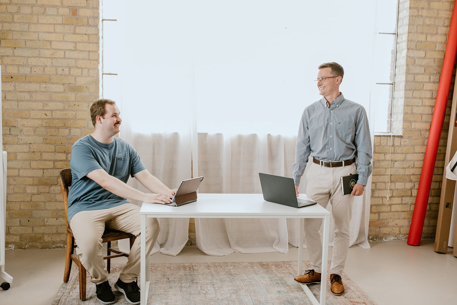 Two men in a bright ECCG office space, one sitting at a desk with a laptop, the other standing with a laptop, both smiling and engaged in conversation.