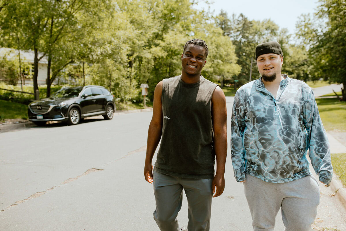 Two men walk side by side on a sunny residential street, smiling at the camera, with trees and a parked car in the background.