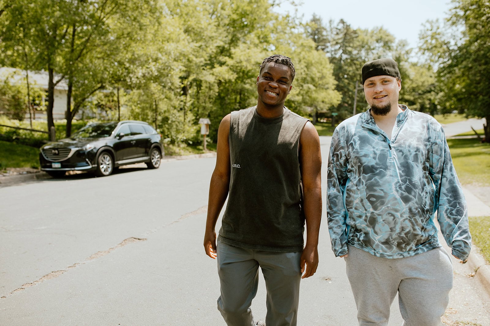 Two men walk side by side on a sunny residential street, smiling at the camera, with trees and a parked car in the background.