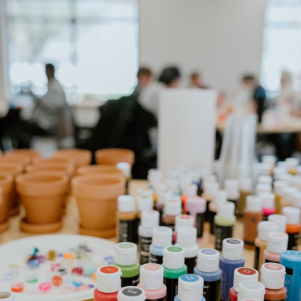 Rows of paint bottles and empty terracotta pots arranged on a table in a bright room, with people sitting and working in the blurred background.
