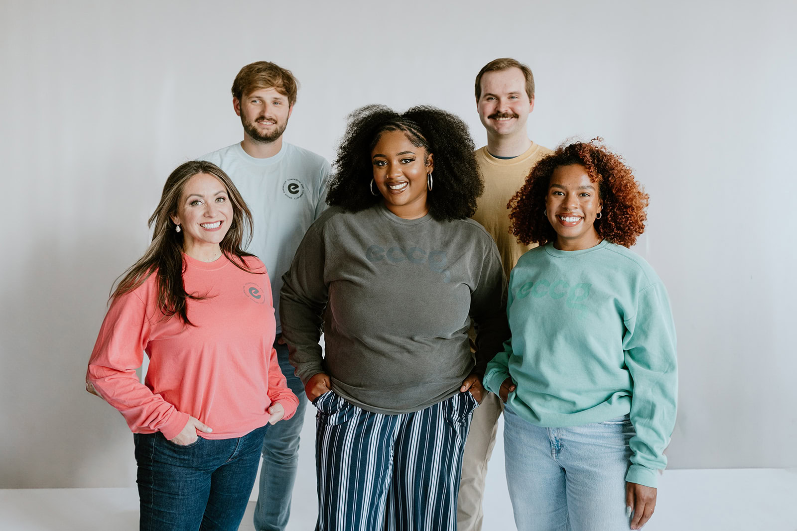 Five people from the ECCG team stand together against a plain background, all smiling and wearing casual long-sleeve shirts in various colors.