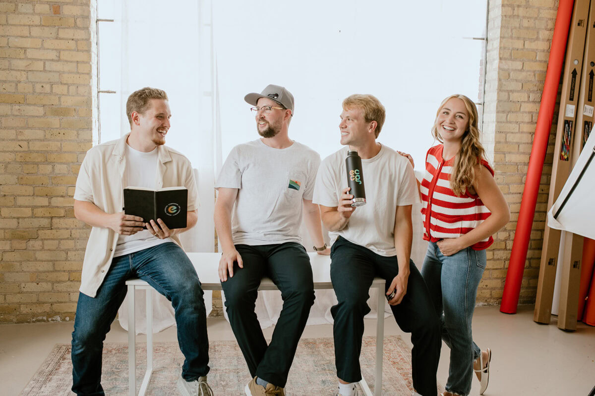 Four people are sitting and standing by a table, smiling and talking. One holds an ECCG notebook, another a water bottle. They are in a room with brick walls and large windows.